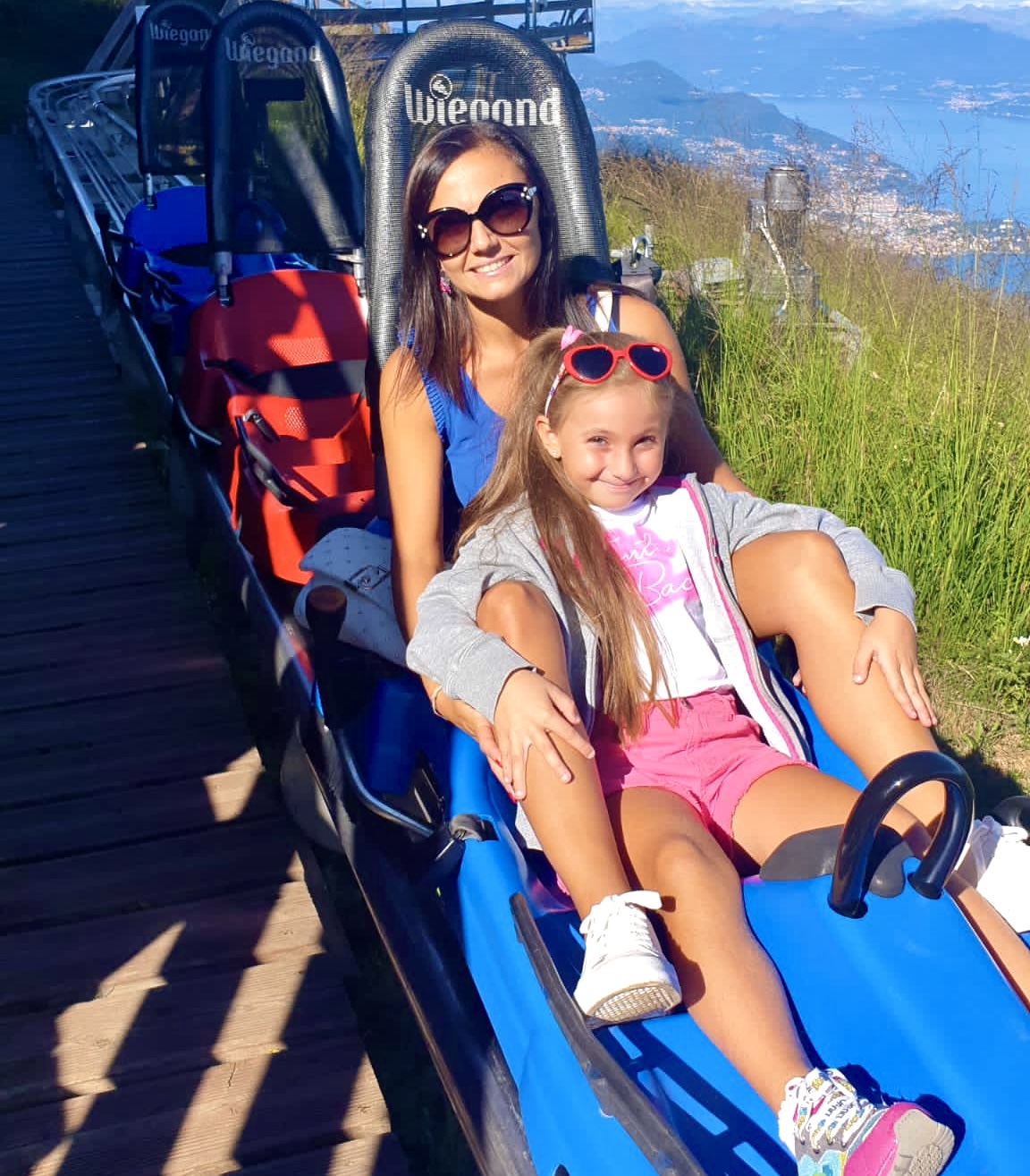 Foto di una mamma con una bambina sedute nel bob di alpyland mottarone nel parco del mottarone con vista sul lago maggiore